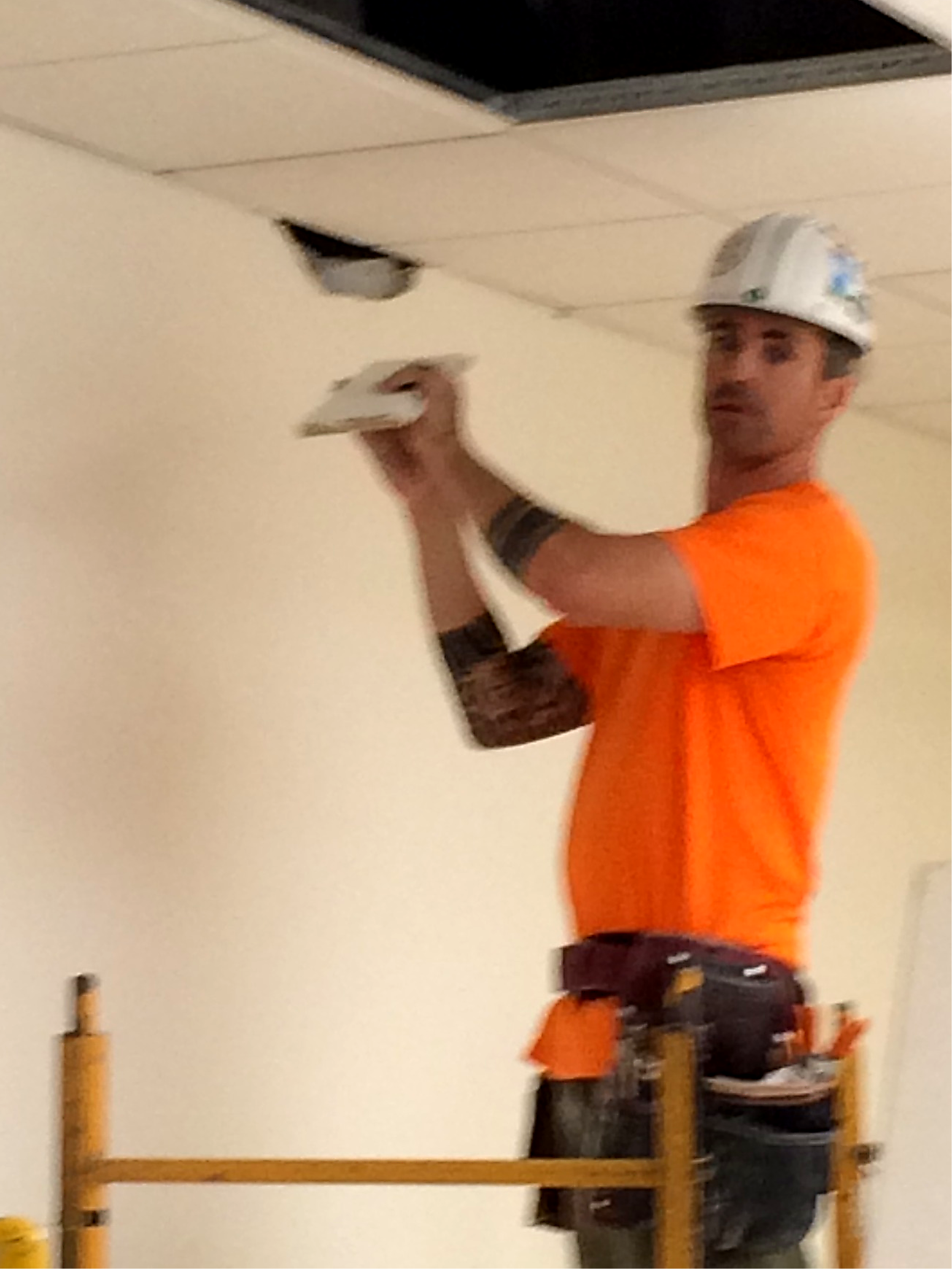 Person in orange shirt and hard hat working on ceiling installation.